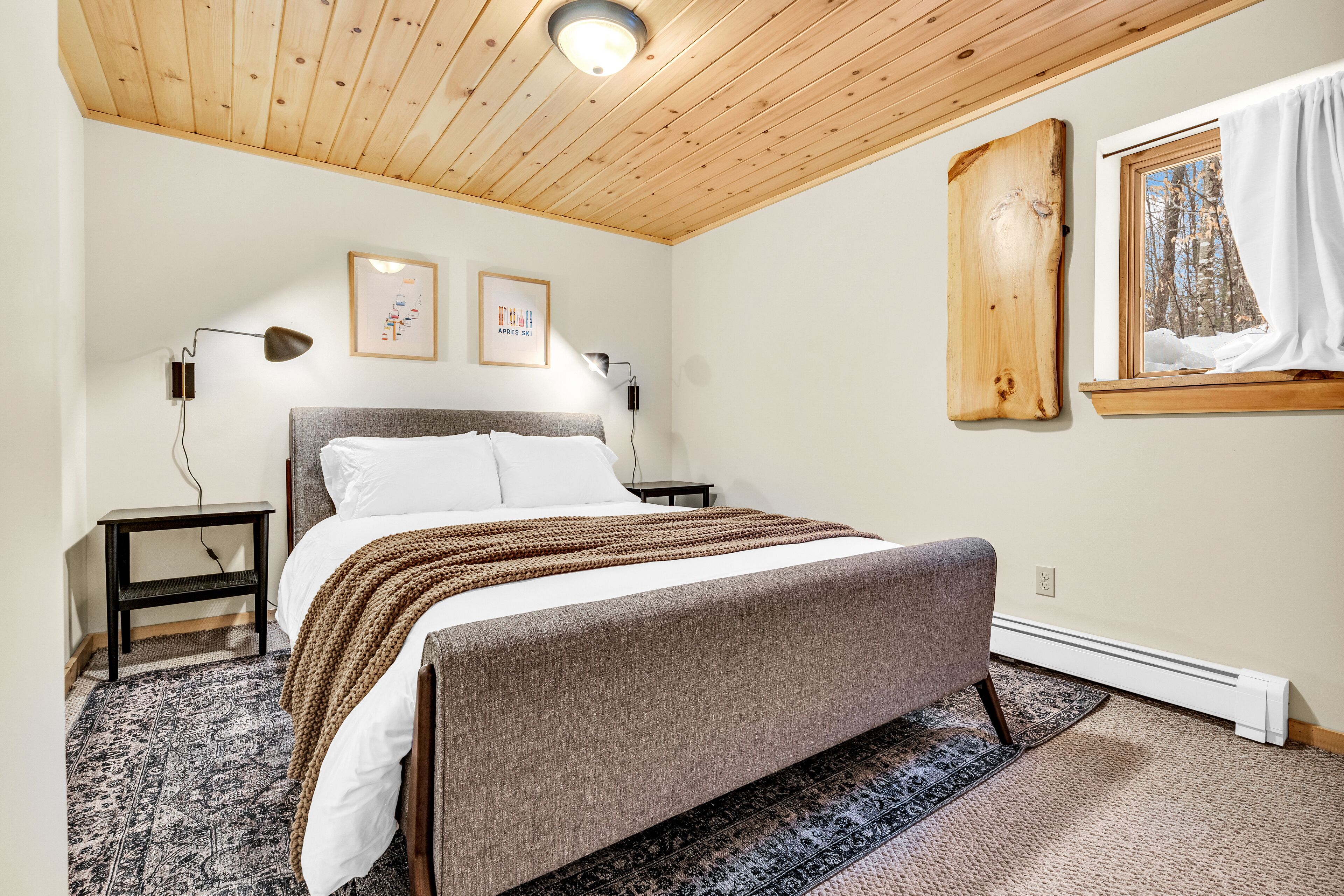 A bedroom with a bed, a nightstand, a wall-mounted heater, and a window with a view of trees.