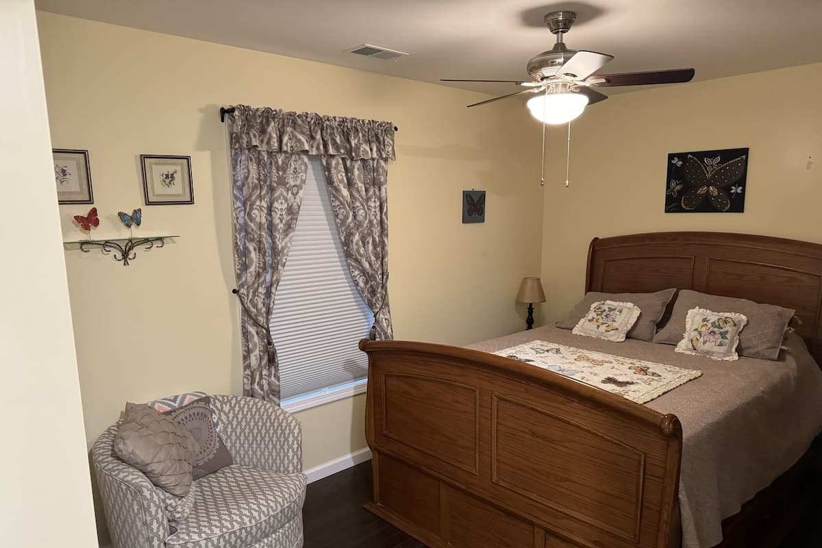 Une chambre à coucher avec un lit en bois, un ventilateur de plafond, une fenêtre avec des rideaux à motifs, une table de chevet et une chaise.