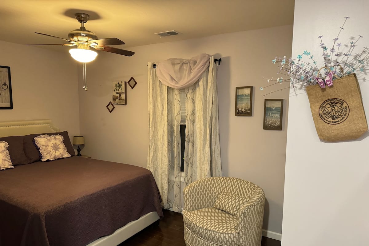 Une chambre à coucher avec un lit, une chaise, un ventilateur de plafond et une fenêtre avec des rideaux.