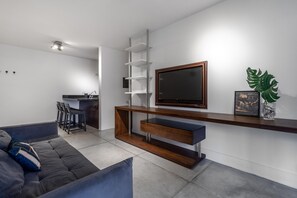 A modern living room with a grey sofa, a wooden entertainment unit, a flat-screen TV, and a small kitchen area with a sink and chairs. - Haddock 166 in S o Paulo (São Paulo)