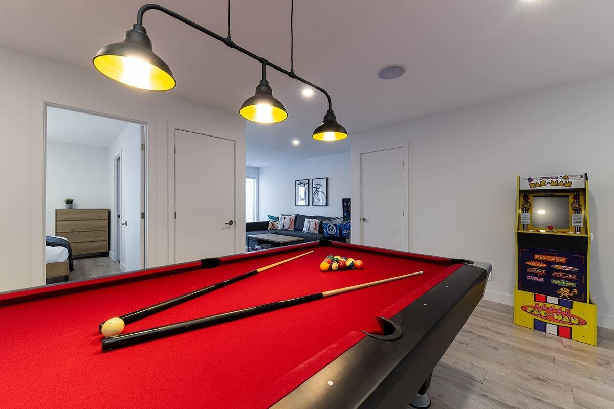 Une table de billard éclairée par trois lampes suspendues, un canapé et une borne d’arcade Pac-Man dans une pièce aux murs blancs, avec une porte donnant sur une autre pièce.
