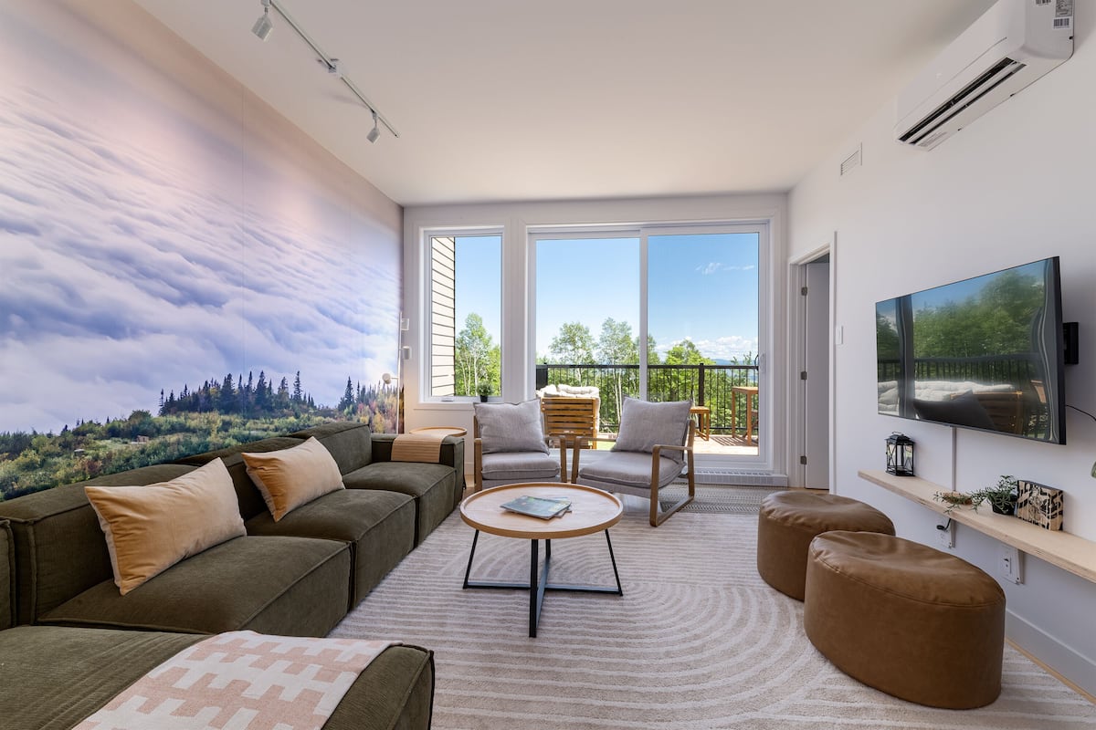 Un salon moderne avec un canapé, des fauteuils, des poufs, une table basse et un téléviseur fixé au mur.