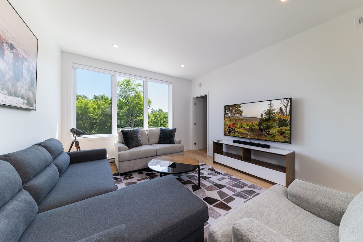 Un salon moderne avec un canapé d’angle gris, un canapé beige, une table basse et un téléviseur posé sur un meuble blanc.