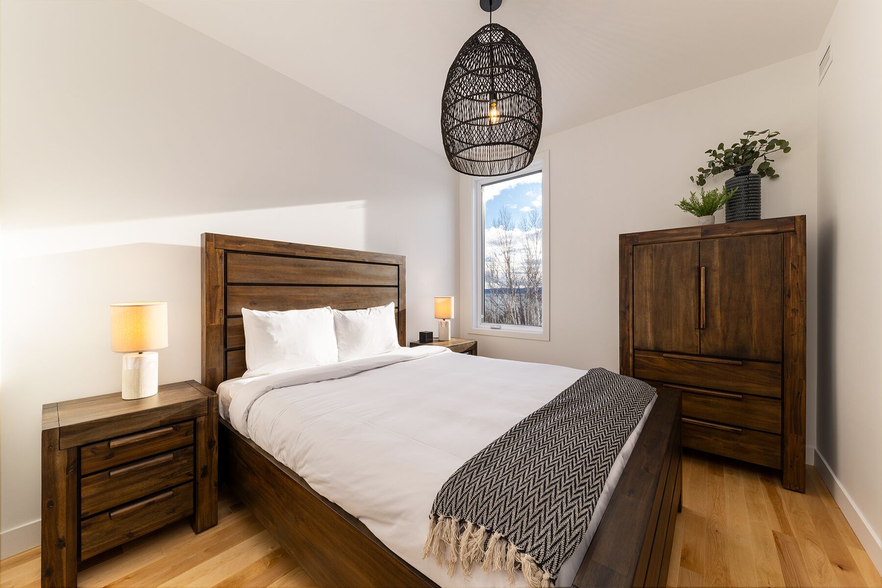 A bedroom with a wooden bed, two bedside tables with lamps, a wardrobe, and a hanging light fixture.