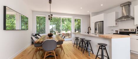 A modern kitchen with a wooden dining table, bar stools, and large windows.