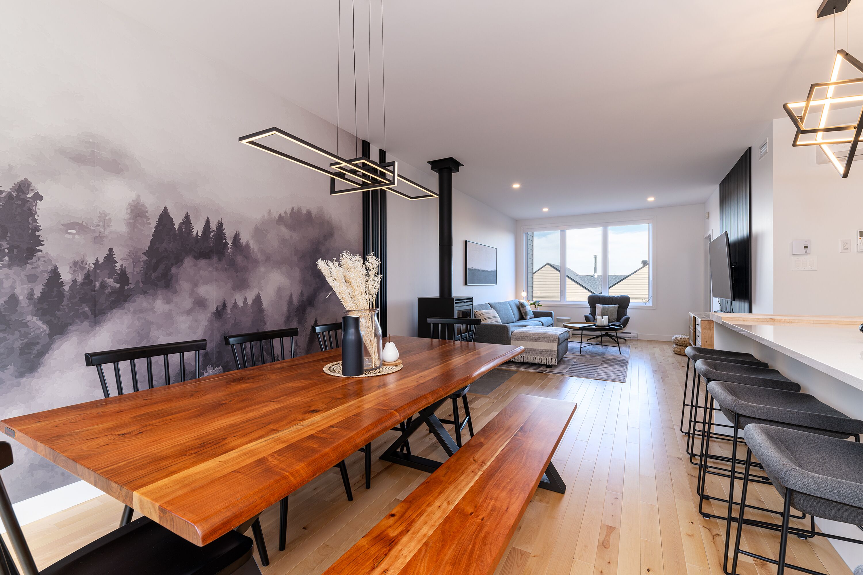 Een moderne eetruimte met een houten tafel, banken en stoelen. Er is een muurschildering, een bloevas en een open haard op de achtergrond.