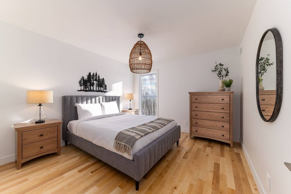 Une chambre à coucher comprenant un lit, des tables de chevet, une commode, un luminaire suspendu et un grand miroir.