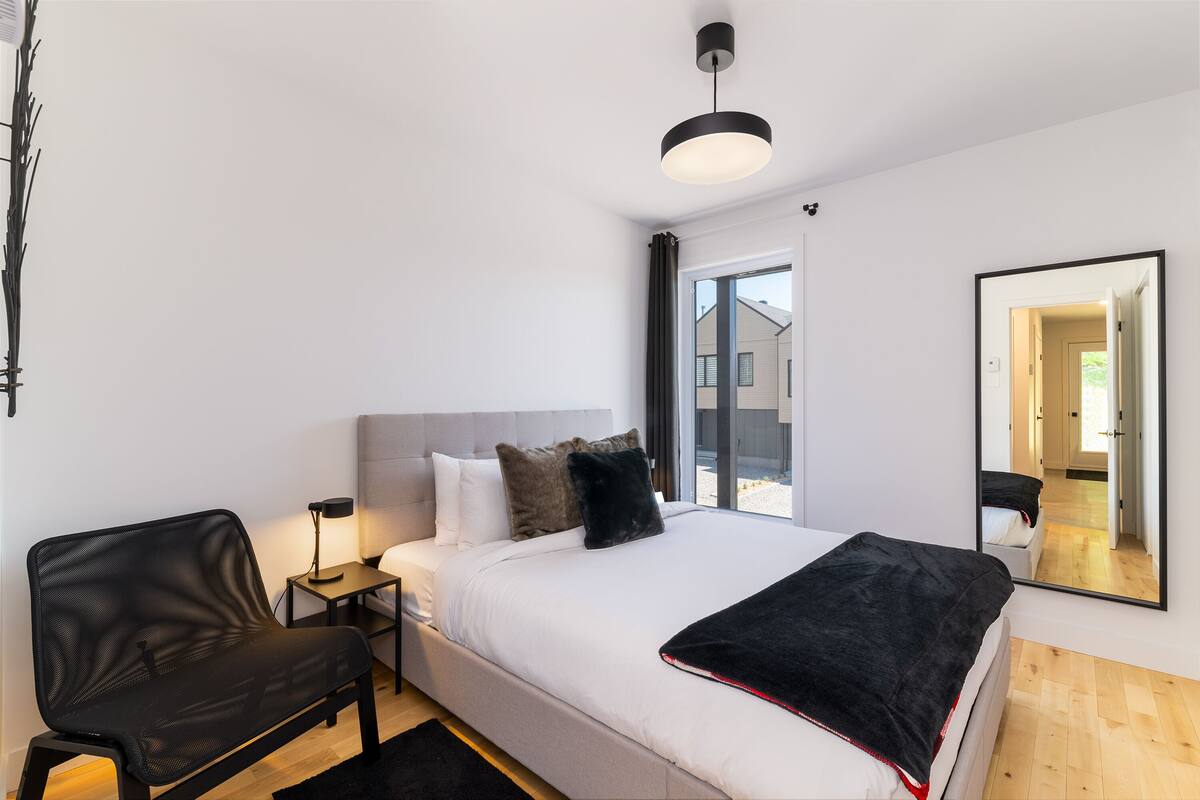 Une chambre à coucher moderne avec un lit, un fauteuil en cuir noir, une table de chevet avec une lampe et un grand miroir au mur.