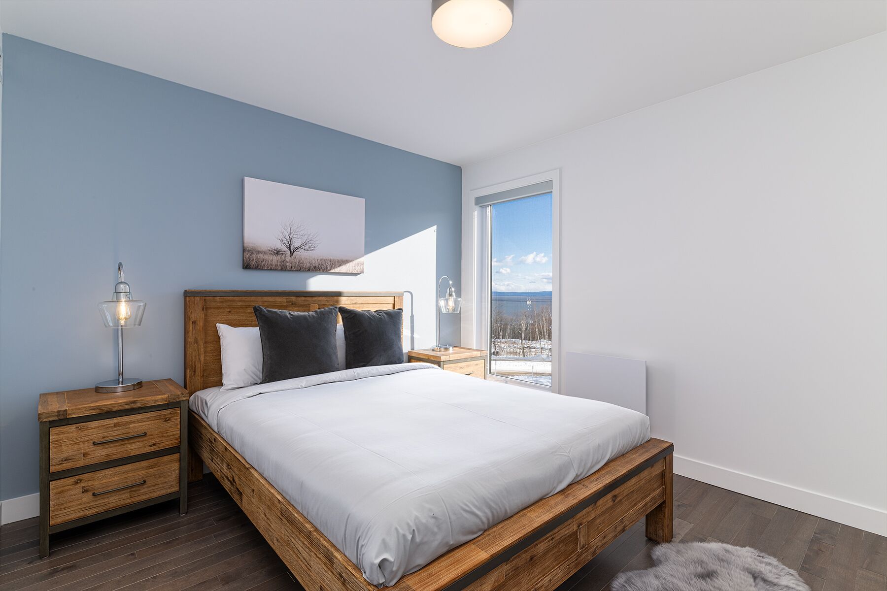 Quarto moderno com cama de madeira, mesa de cabeceira, luminária e uma grande janela com vista para o exterior.