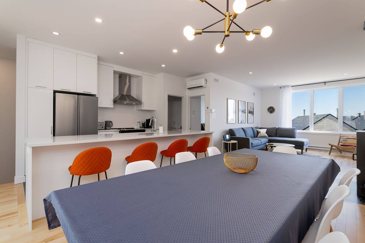 Une cuisine moderne avec des meubles blancs, un réfrigérateur en acier inoxydable et un coin repas agrémenté d’une nappe bleue et de chaises orange.