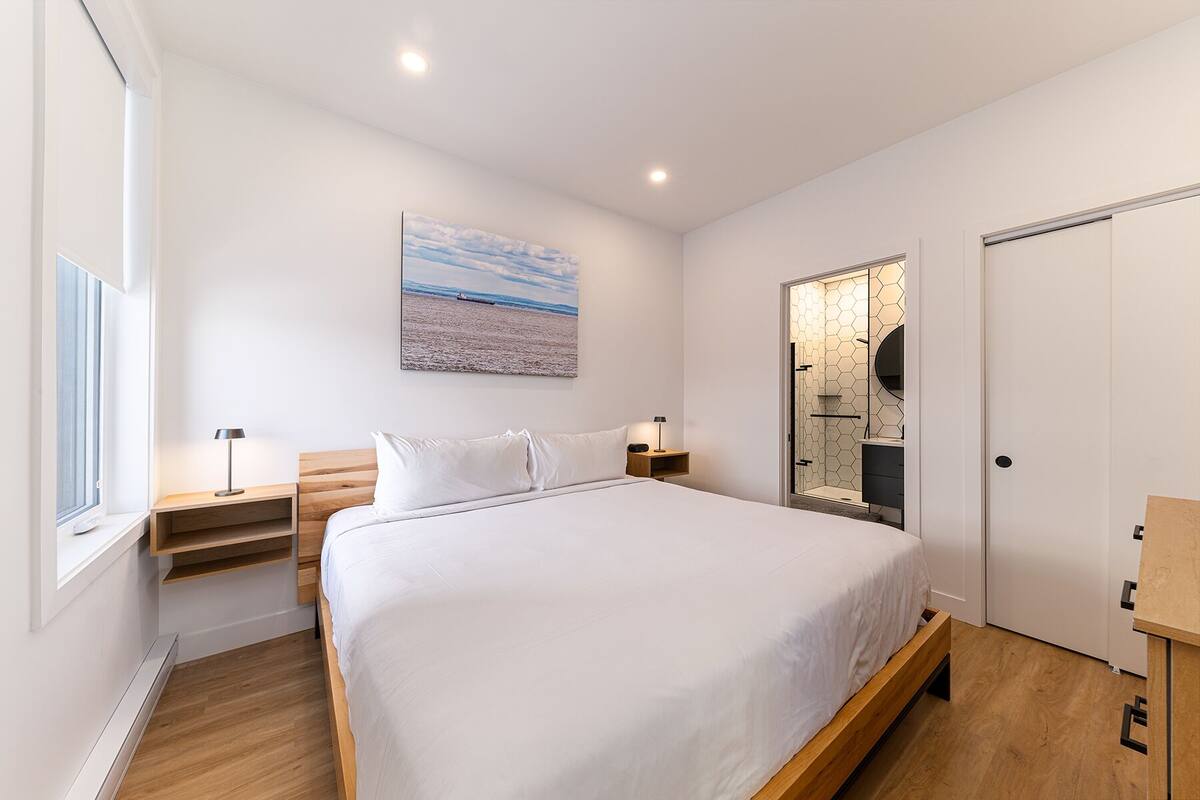 Une chambre moderne avec un grand lit, une table de chevet, une lampe, un tableau représentant une plage et une salle de bain avec un revêtement mural en carreaux hexagonaux.