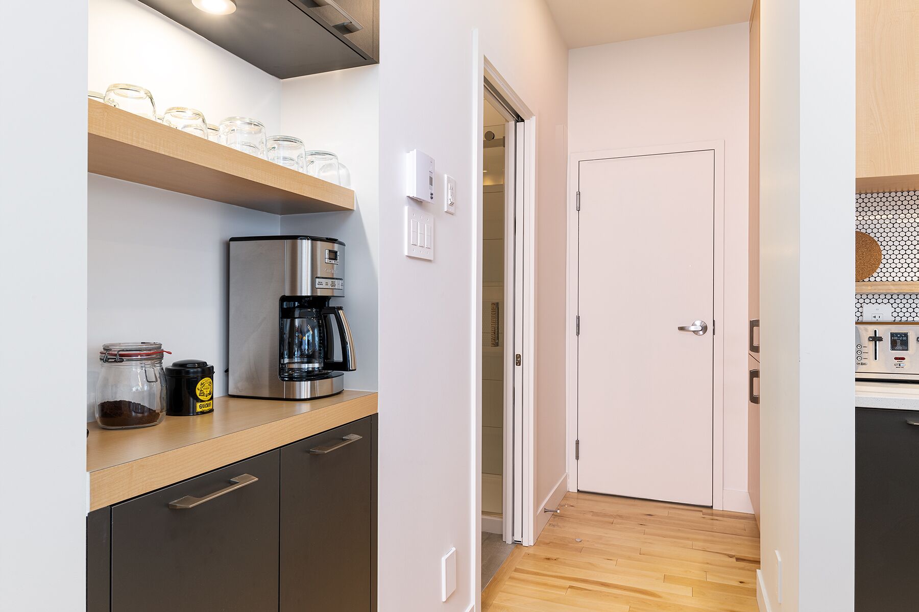 A compact kitchen with a coffee maker, jars, and a door leading to another room.