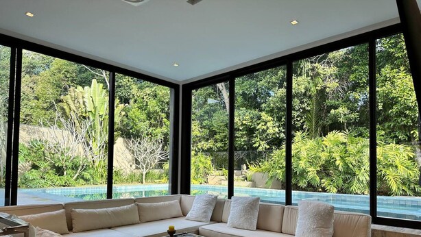 Ein modernes Wohnzimmer mit einem großen Ecksofa, einem Couchtisch und Blick auf einen Swimmingpool sowie üppiges Grün durch große Fenster.