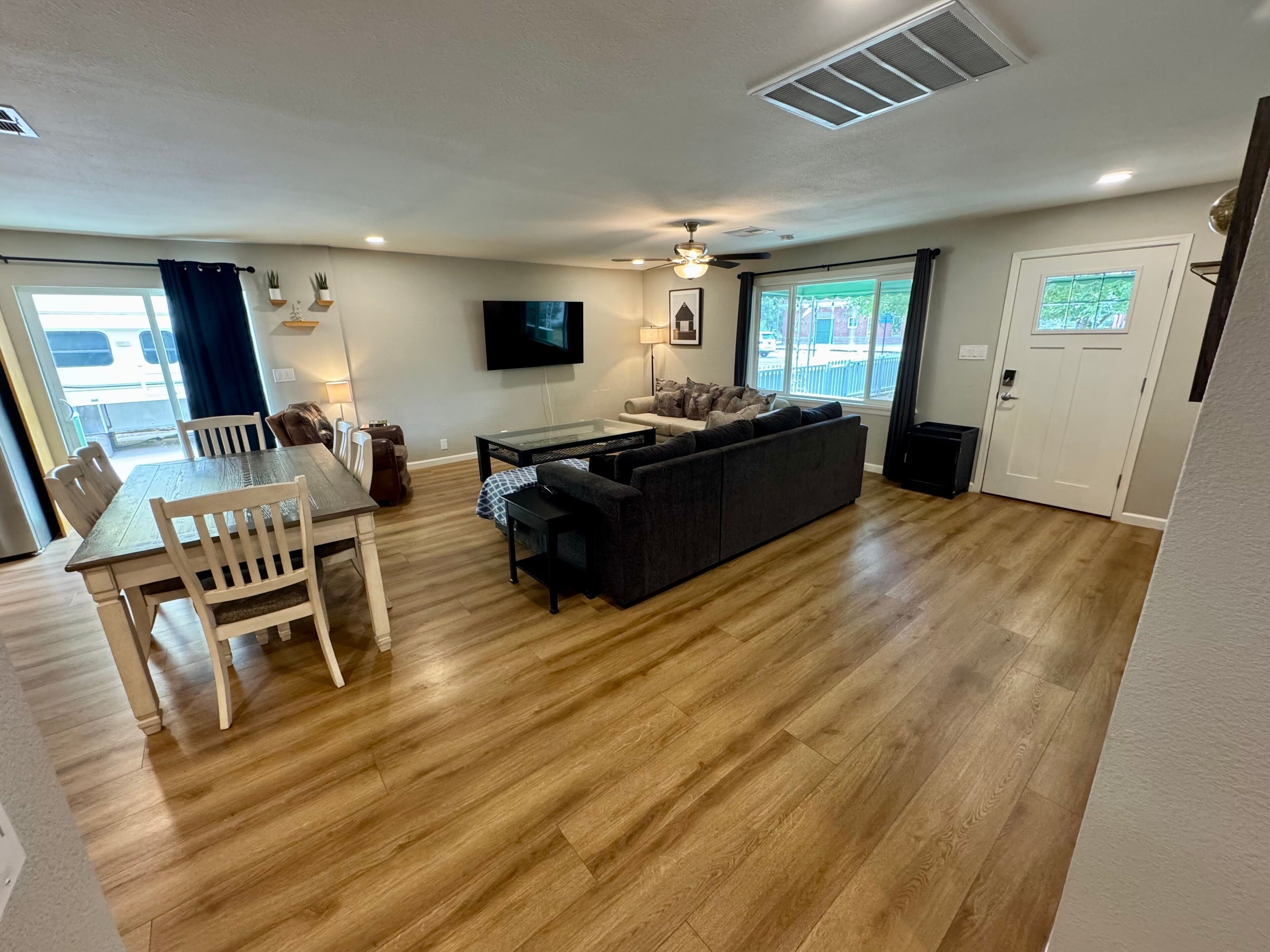 A spacious living room with a wooden floor, a dark-colored sofa, a glass-top coffee table, and a flat-screen TV mounted on the wall.