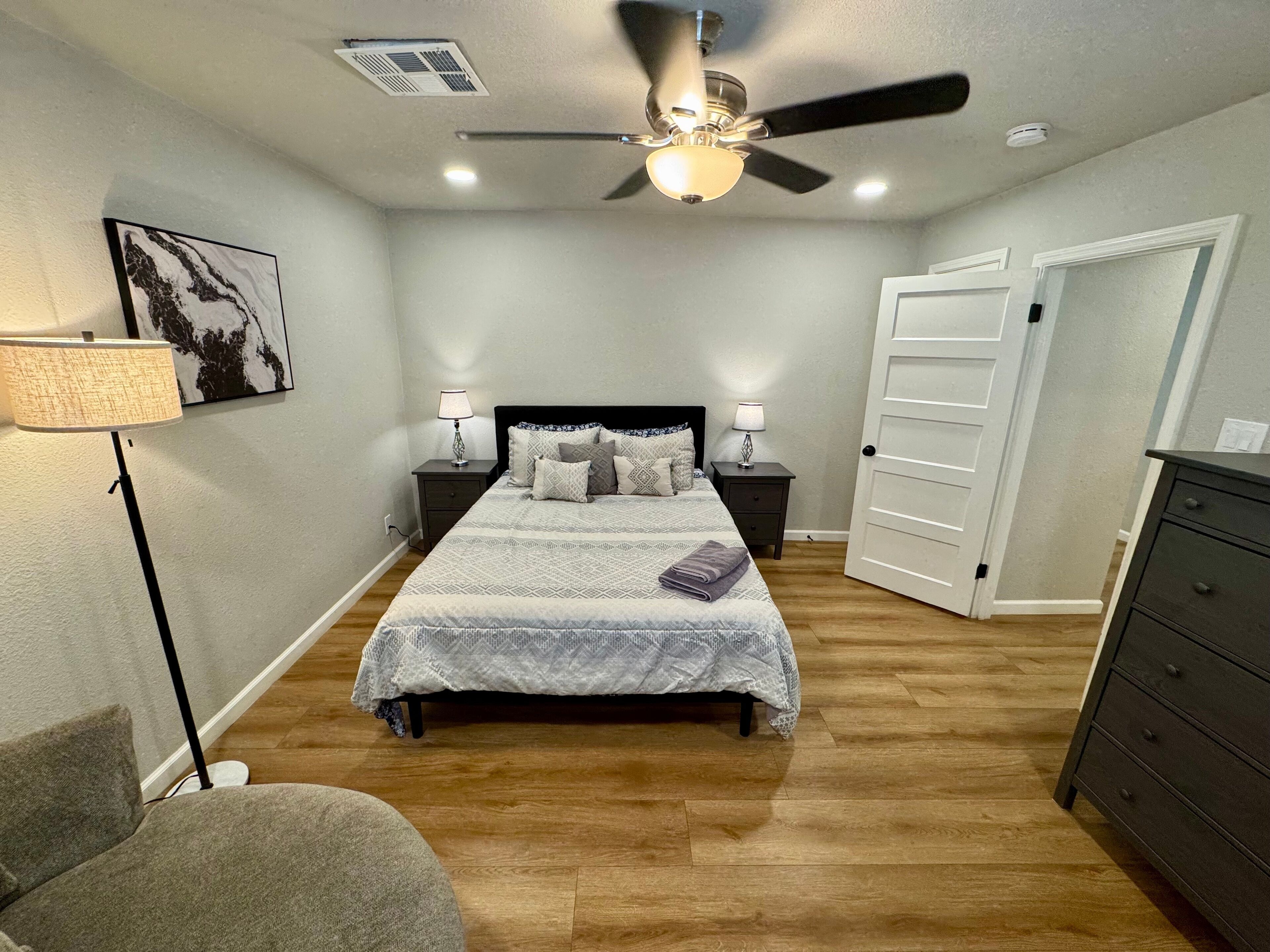A bedroom with a bed, two bedside tables with lamps, a ceiling fan, a door, and a dresser.