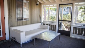 Katettu kuisti, jossa on valkoinen sohva ja metallipöytä, puuseinät ja ikkunoista avautuva näkymä siniseen rakennukseen.