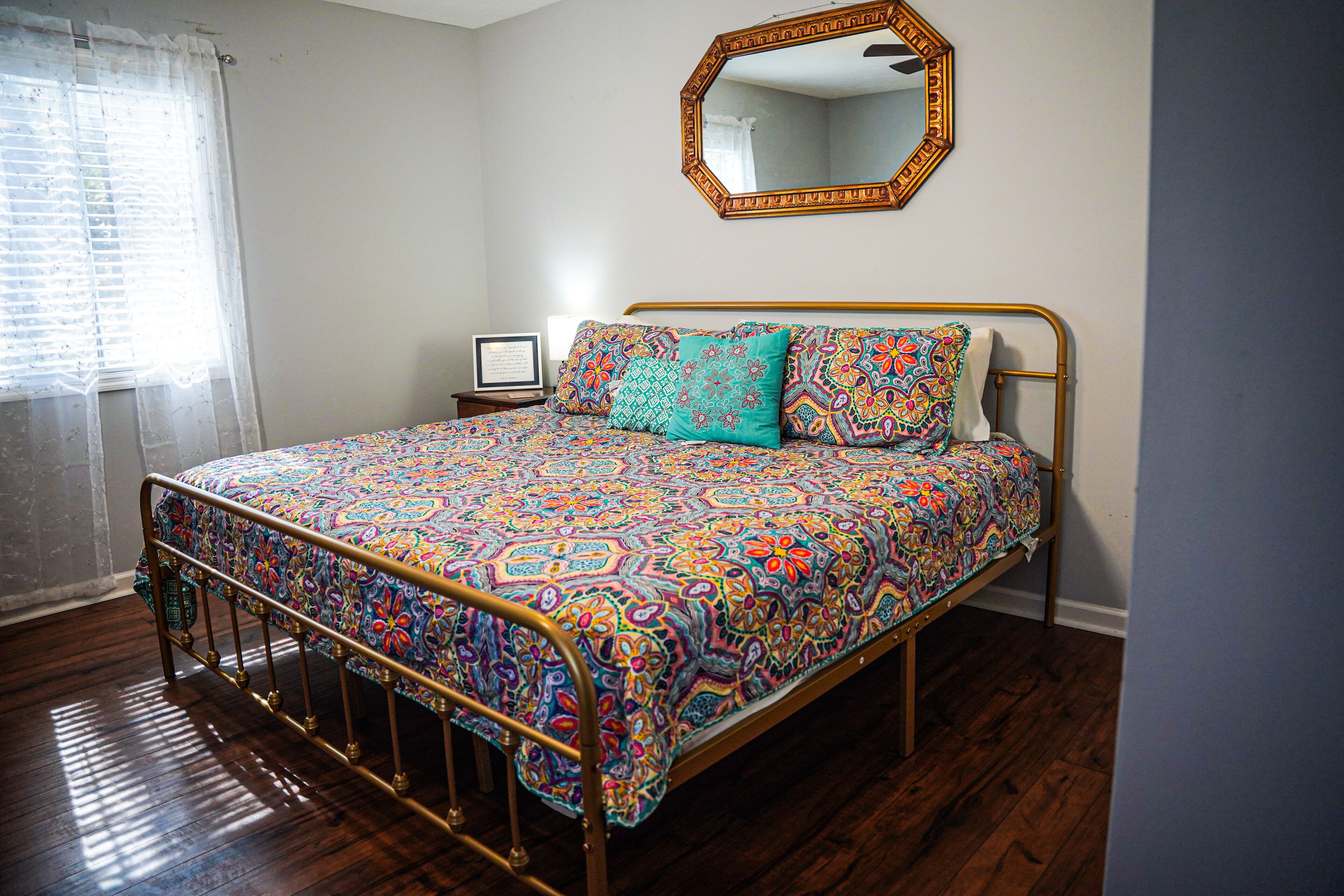 A bedroom with a metal bed frame, patterned bedding, a bedside table with a lamp, and a mirror on the wall.