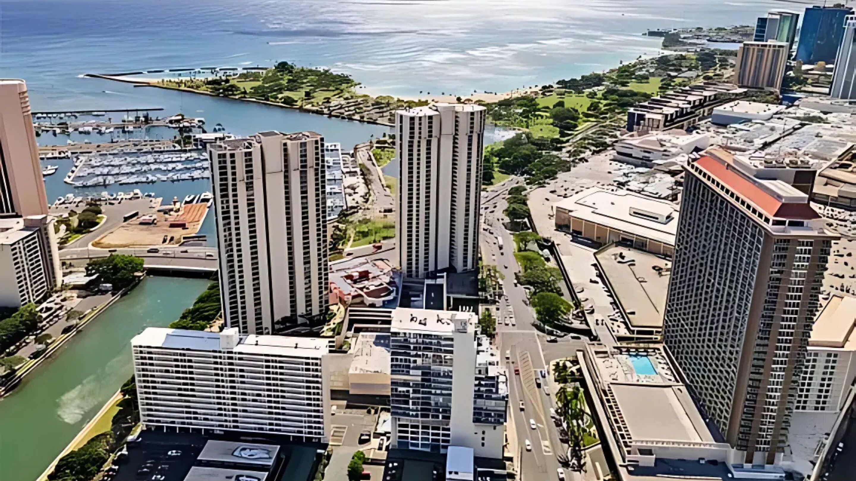 Um edifício alto com design moderno, rodeado por outros edifícios altos e uma marina com barcos.