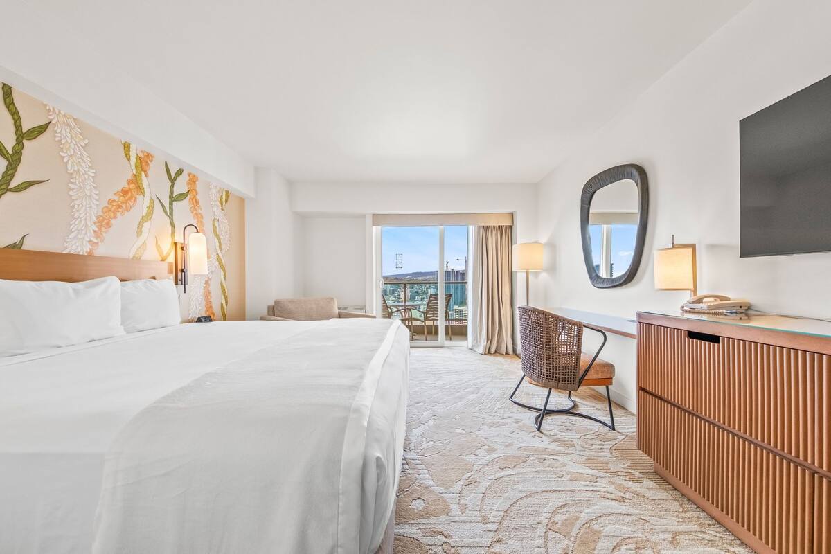 Ein Hotelzimmer mit einem großen Bett, einem Schreibtisch mit Telefon, einem Sessel und einem großen Spiegel sowie Blick auf die Stadt durch das Fenster.
