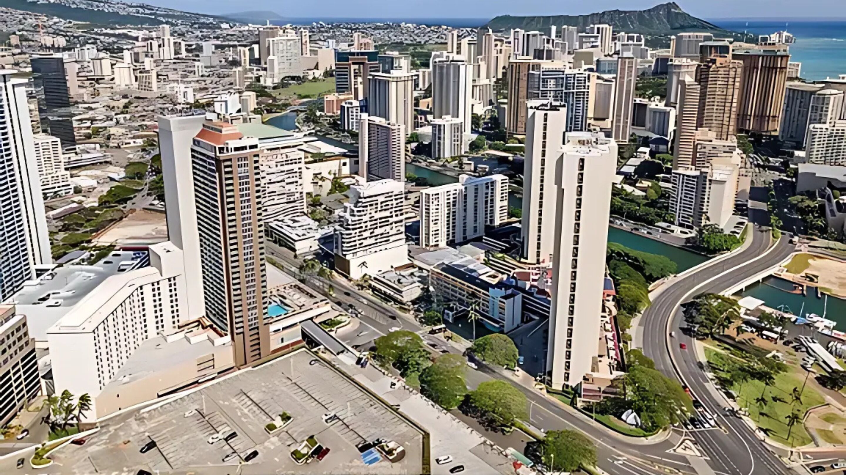 Área urbana com edifícios altos e arquitetura moderna, uma via central e um corpo d'água ao fundo.