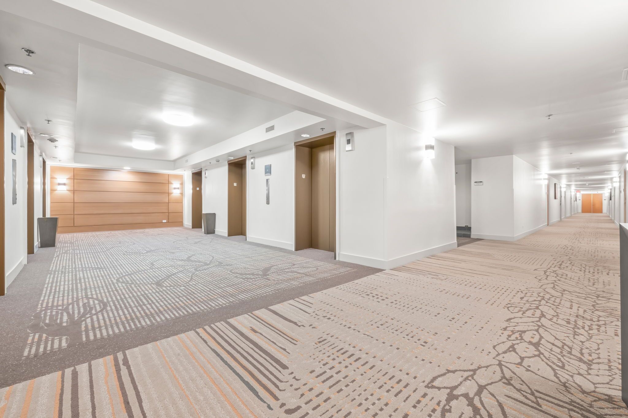 Um corredor longo e bem iluminado, com várias portas, piso coberto de carpete e iluminação embutida.