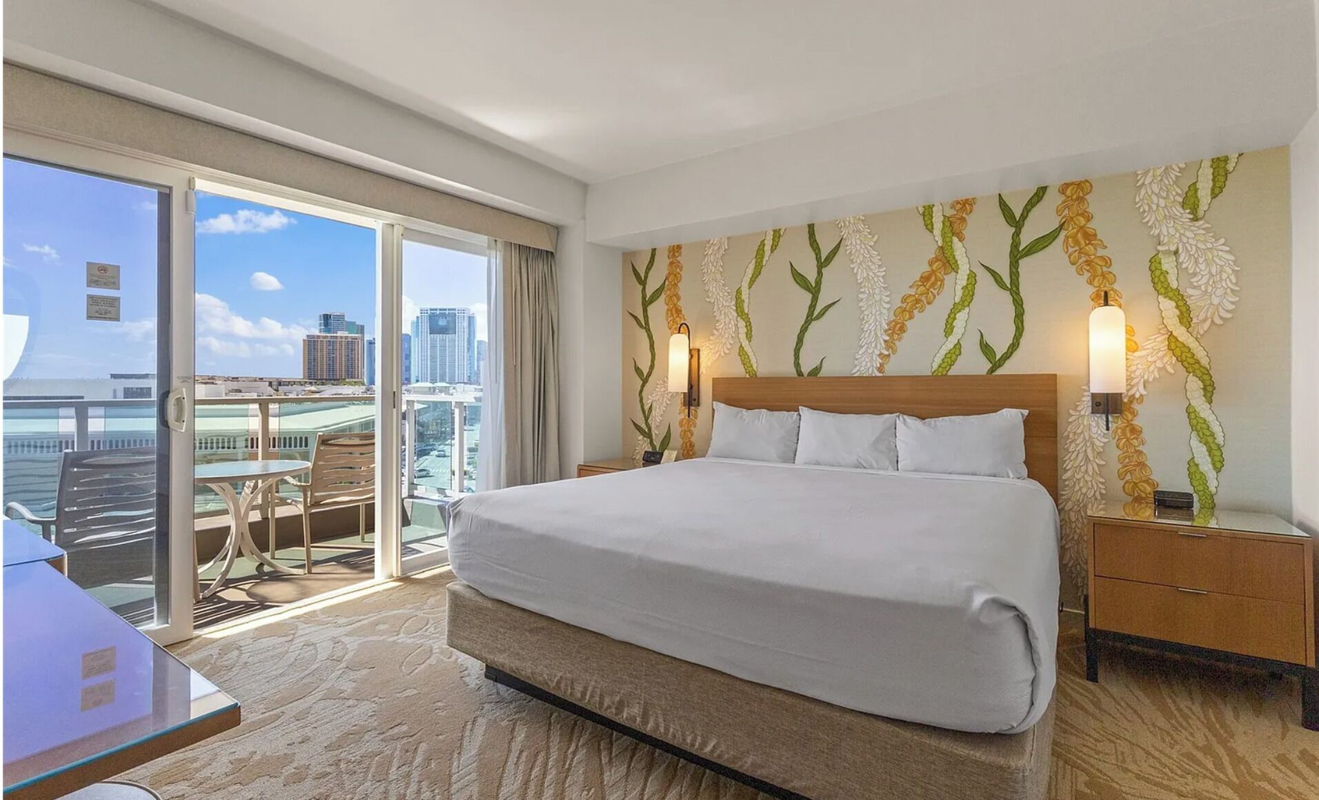 A hotel room with a large bed, a desk, and a balcony with a city view.