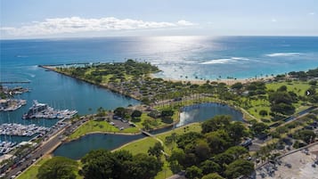 An aerial view of a coastal area with a marina, a large body of water, and a park with pathways and greenery.