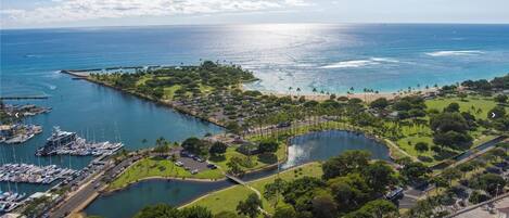 An aerial view of a coastal area with a marina, a large body of water, and a park with pathways and greenery.
