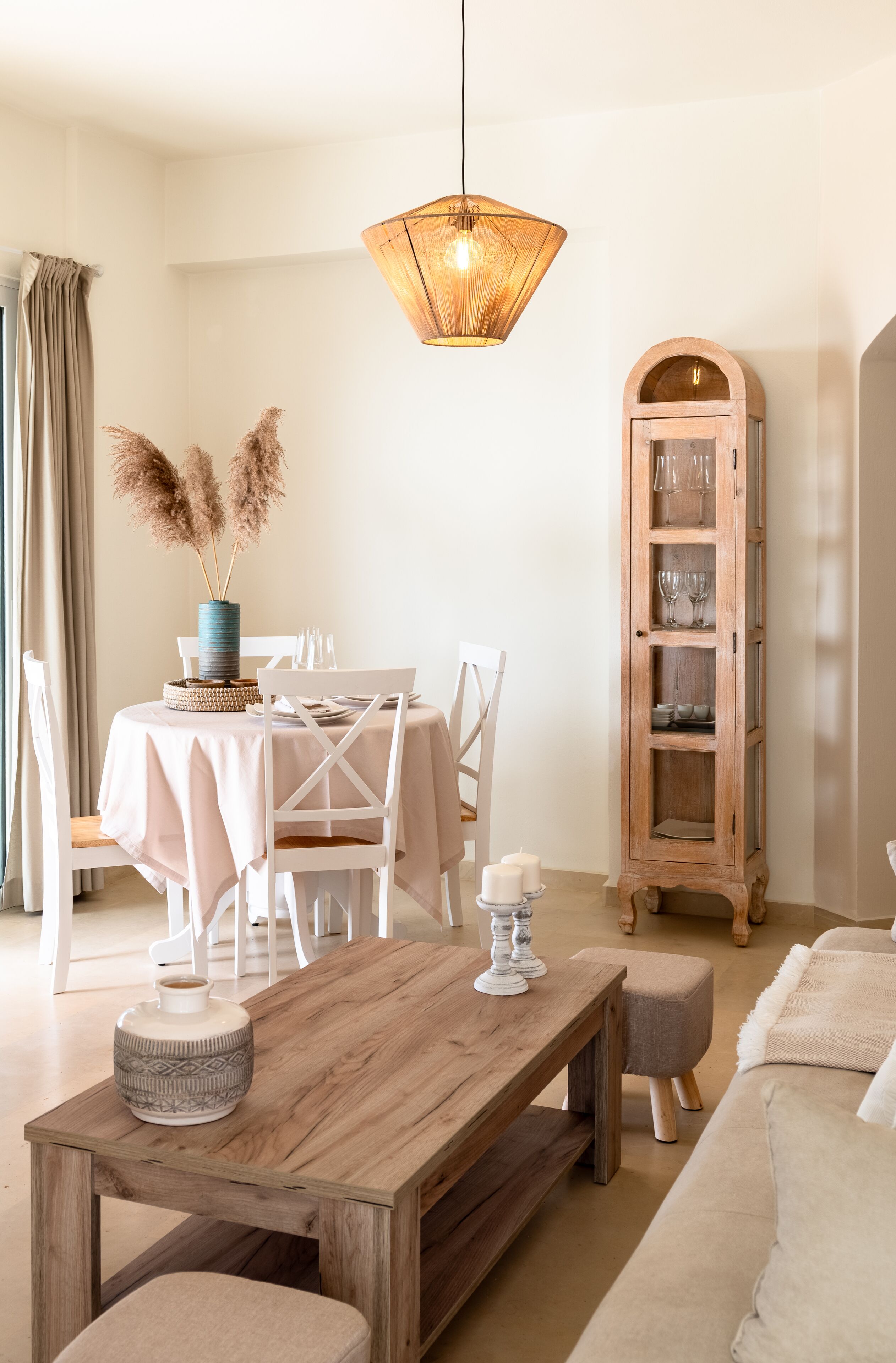 A dining area with a wooden table, white chairs, a hanging light fixture, and a tall wooden cabinet with glassware.