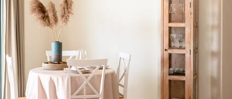 A dining area with a wooden table, white chairs, a hanging light fixture, and a tall wooden cabinet with glassware.
