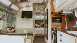 Ein kompaktes Wohnmobil-Interieur mit Küchenzeile, einer kleinen Essecke, einem Bett mit Fernseher und einem Fenster mit Pflanzen.