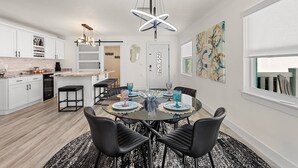 A modern kitchen with white cabinets, a dining area with a glass table, black chairs, and a unique ceiling light fixture. - Waterfront! 1BR with private pool! 6 min. walk to the beach! (Treasure Island)