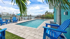 A swimming pool with blue chairs, a dock with boats, and a view of the water and buildings in the background. - Waterfront! 1BR with private pool! 6 min. walk to the beach! (Treasure Island)