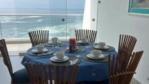 A dining table with a blue tablecloth, surrounded by wicker chairs, set for four people with plates, cups, and a glass pitcher. A large window offers a view of the ocean. - Terrace with Jacuzzi, Duplex, Ocean View, Grill (Punta Hermosa)