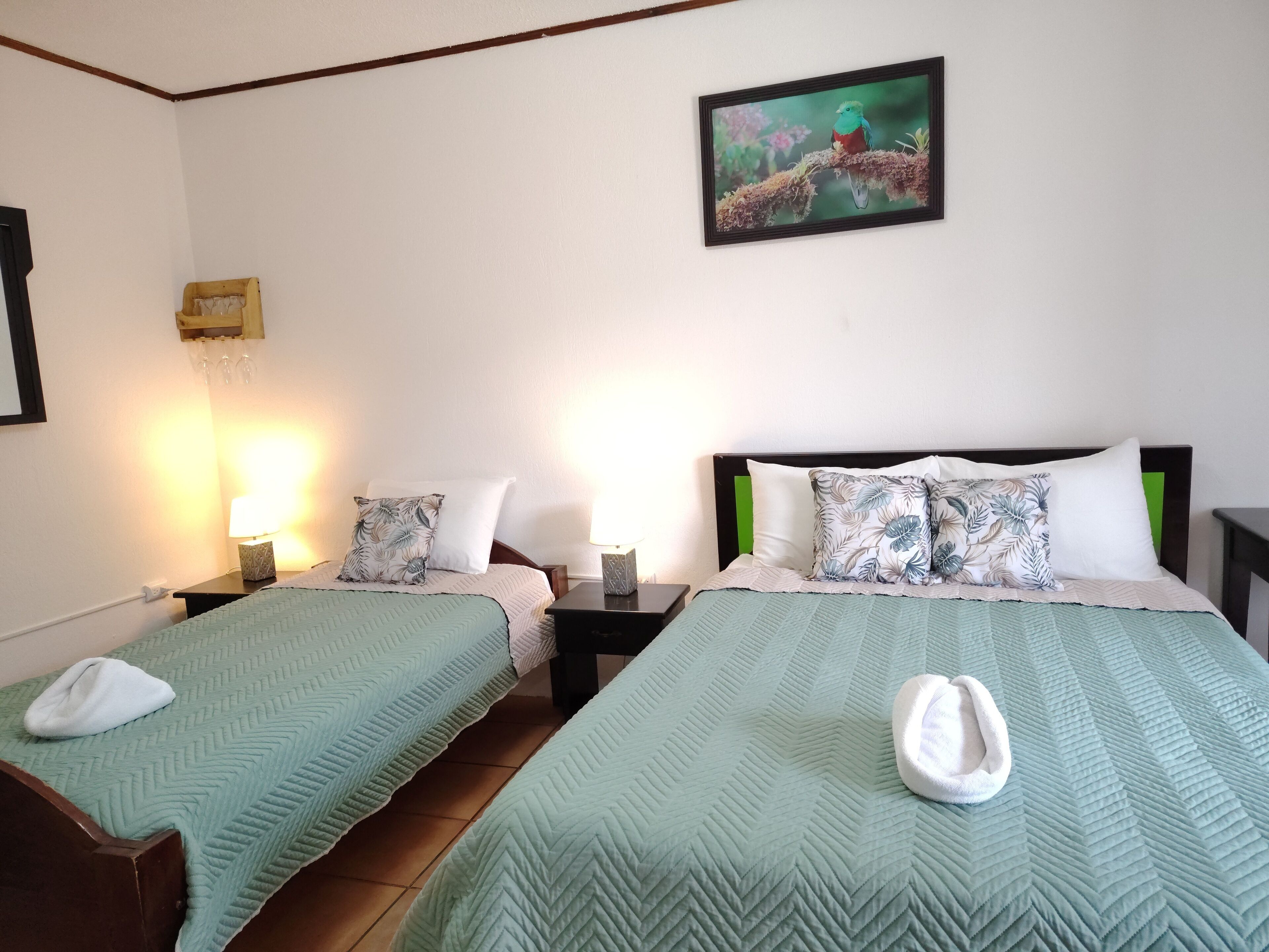 A hotel room with two beds, each with a green quilt, white pillows, and a wooden headboard. There is a painting of a bird on the wall, a small table with a lamp, and a wall-mounted shelf with glasses.