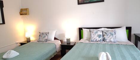 A hotel room with two beds, each with a green quilt, white pillows, and a wooden headboard. There is a painting of a bird on the wall, a small table with a lamp, and a wall-mounted shelf with glasses.