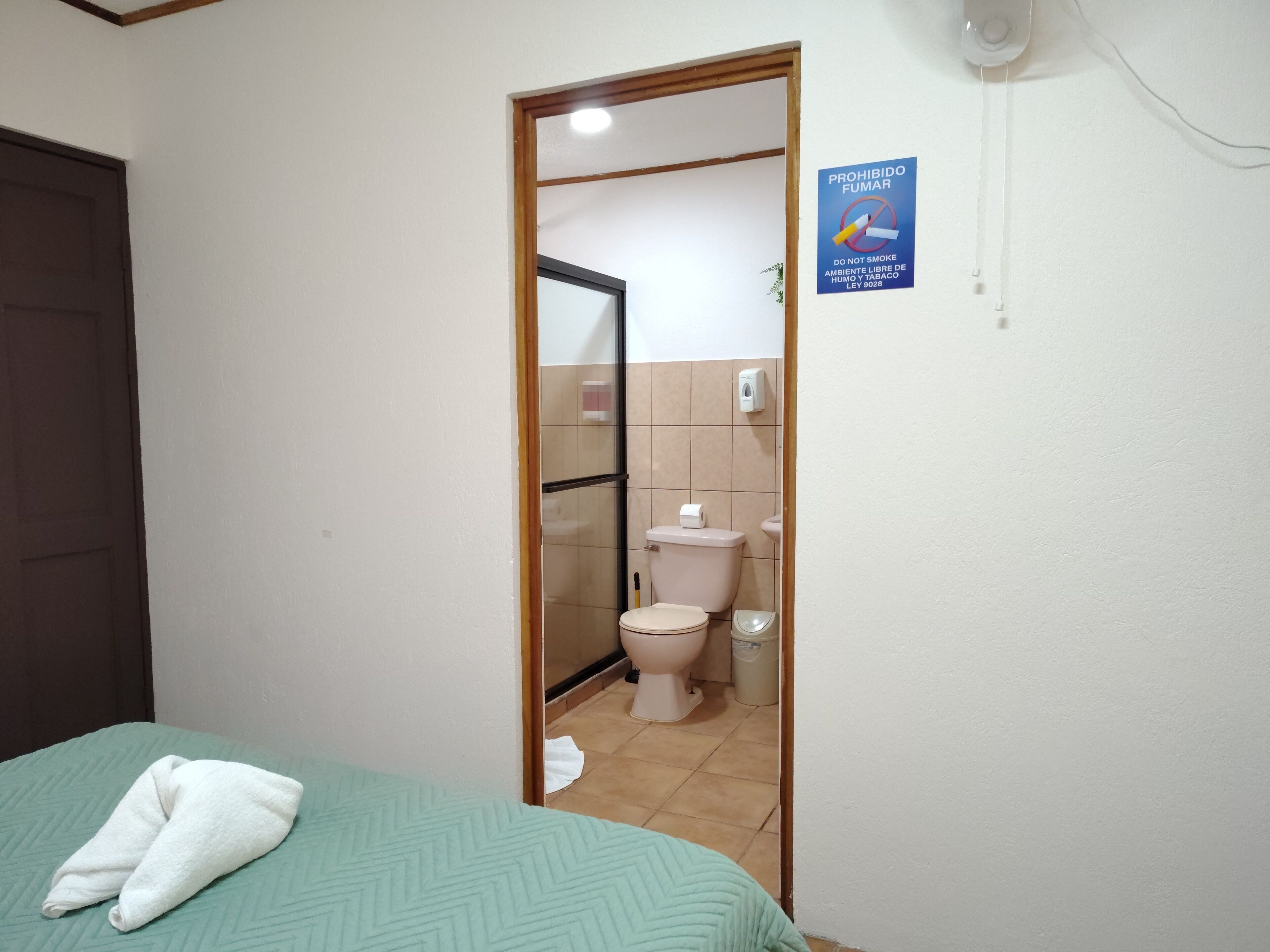 A hotel room with a bed, a bathroom visible through an open door, and a no-smoking sign on the wall.