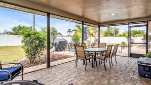 An outdoor patio with a table and chairs, a grill, and a view of a backyard with a pool and palm trees. - Perfect for Families, Friends and Budget of all Sizes! Great Deal! Heated Pool! (Cape Coral)