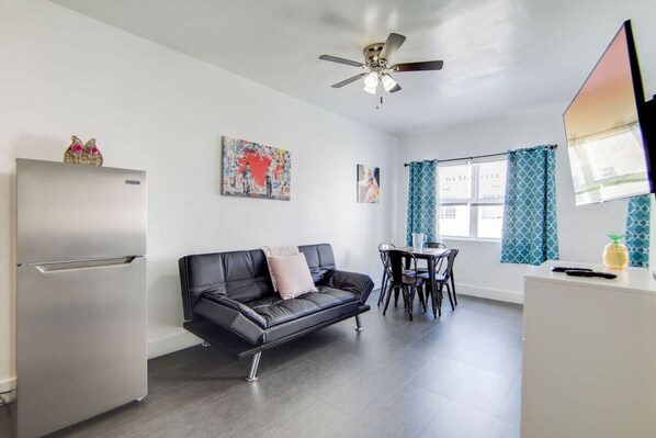 A compact living space with a kitchenette, a black leather sofa, a ceiling fan, and artwork on the walls.