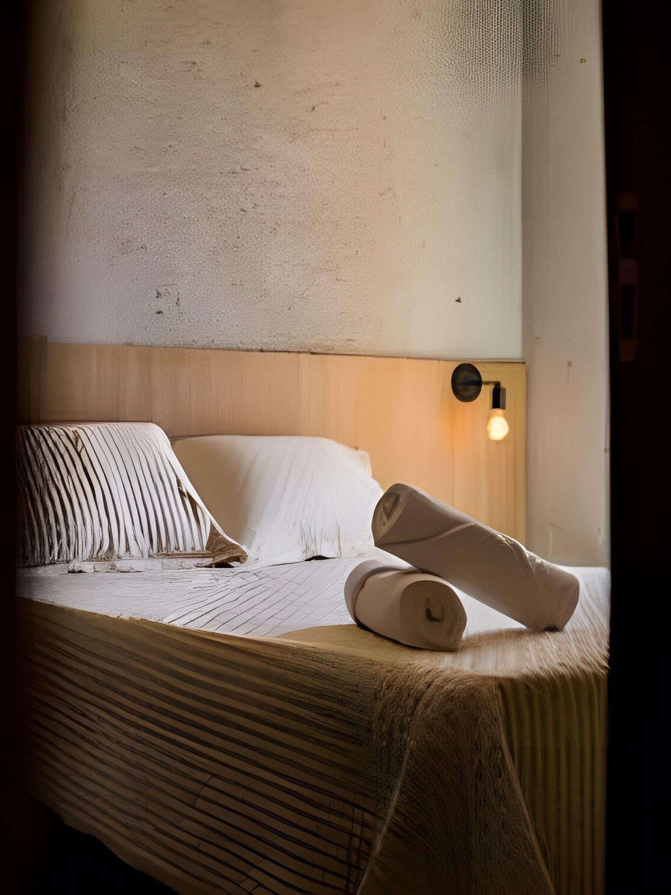 Uma cama bem arrumada com lençóis brancos, uma cabeceira de madeira e uma luminária fixada na parede.