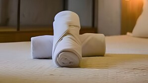 A neatly made bed with rolled towels, a wooden headboard, and a wall-mounted light fixture.