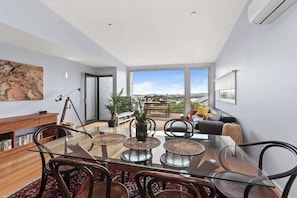 A modern living room with a glass dining table, comfortable seating, a large window with a city view, and a bookshelf. - Marriners Lookout Retreat (Apollo Bay)