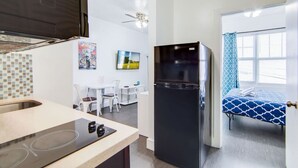 A compact kitchen with a black refrigerator, a dining area with white chairs, and a bed with a blue patterned bedspread. - Comfy 1 B/R, Near Ocean Dr & Beach - South Beach (Miami Beach)