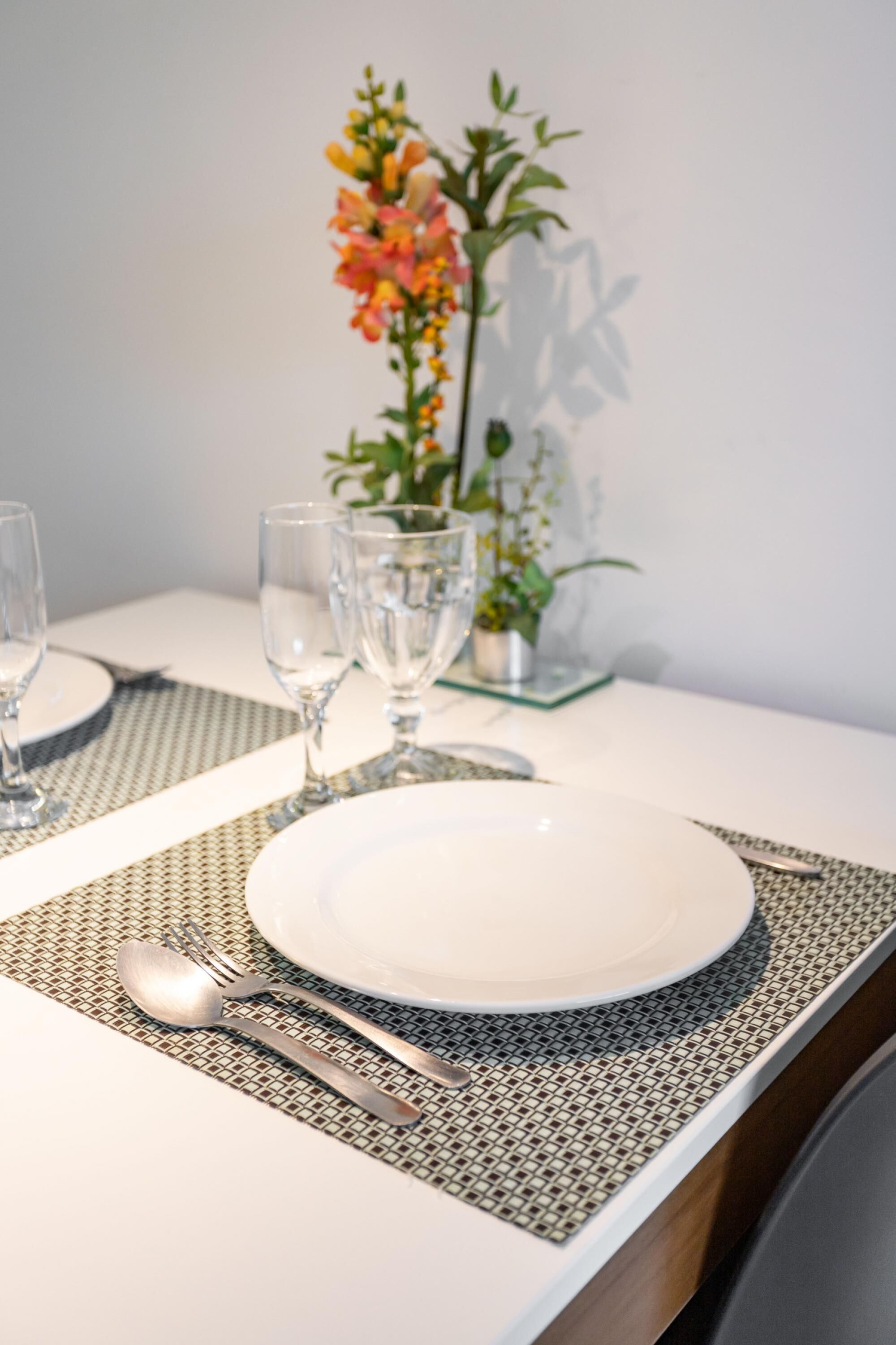 A neatly set dining table with a white plate, silverware, and clear glasses, accompanied by a decorative plant in the background.