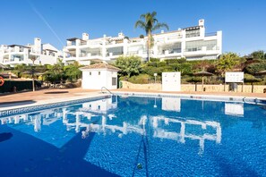 A swimming pool with clear blue water, surrounded by a white building with balconies and a small garden area.