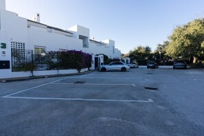 A parking lot with several cars, a white building with a number '3' sign, and a small tree with purple flowers. - El Bosque de la Mairena (Ojen)