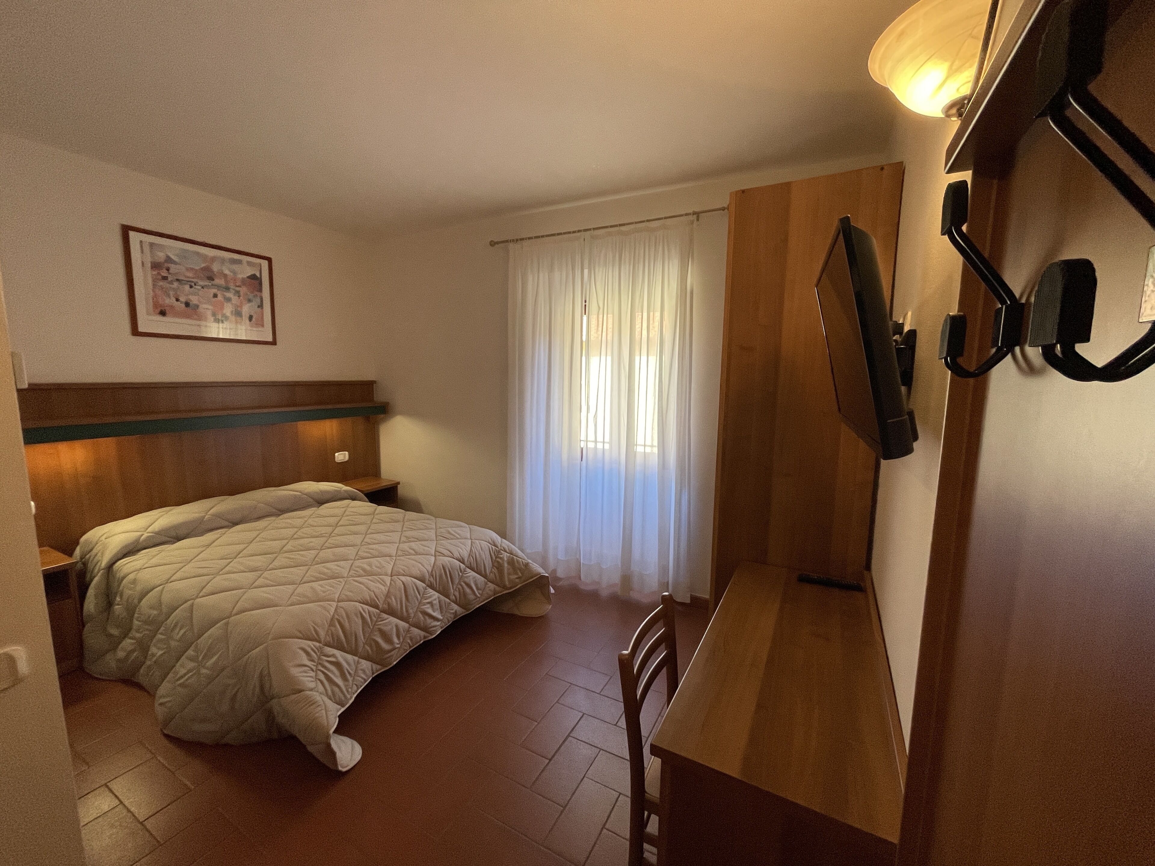 A hotel room with a bed, a desk, a chair, a window with curtains, a wall-mounted lamp, and a framed picture on the wall.