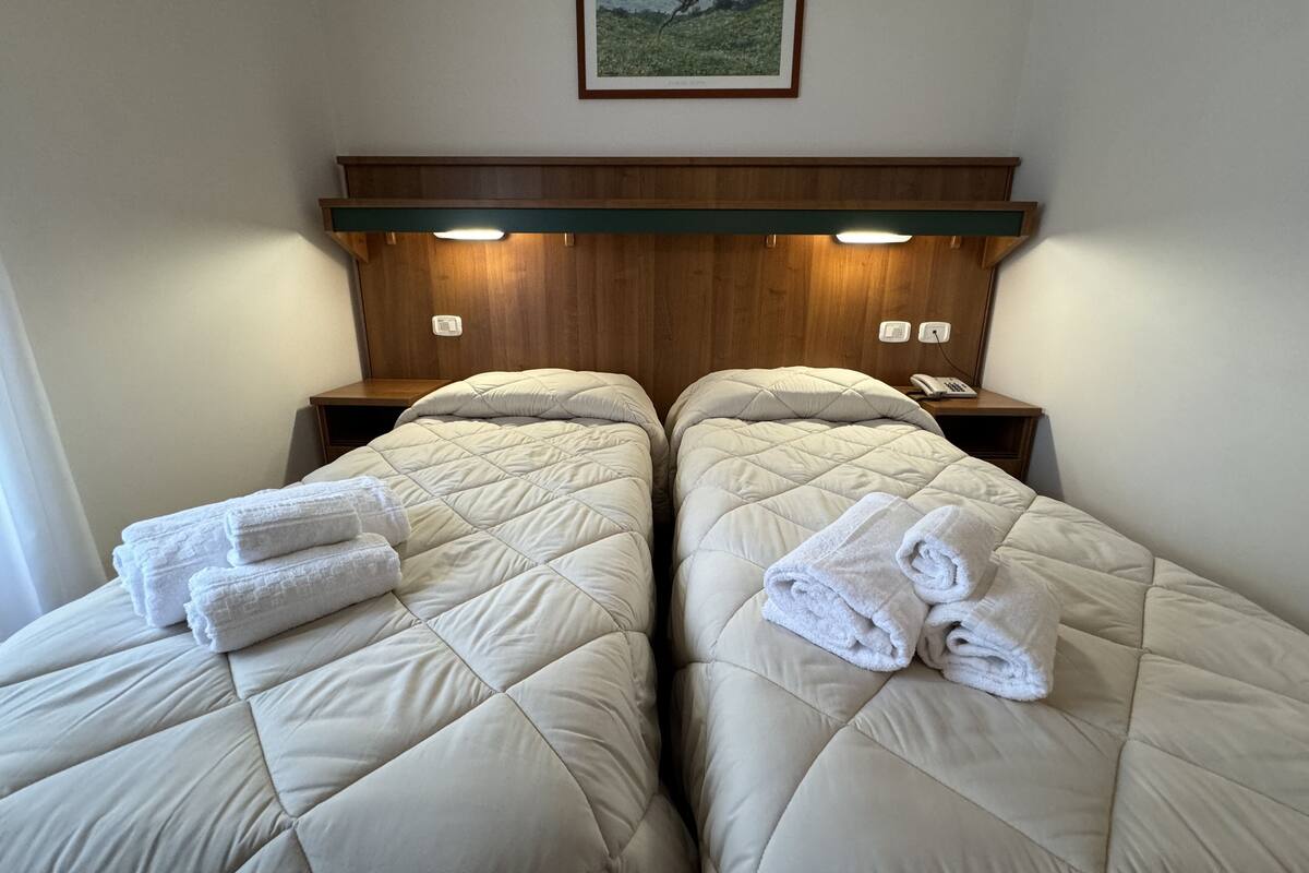 Une chambre d’hôtel avec deux lits, chacun doté d’un matelas matelassé et d’une serviette pliée. On y trouve une tête de lit en bois avec un éclairage intégré, une table de chevet avec un téléphone et un tableau encadré au mur.
