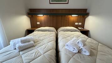 A hotel room with two beds, each with a quilted mattress and a folded towel. There is a wooden headboard with built-in lighting, a bedside table with a phone, and a framed picture on the wall.