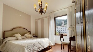 A hotel room with a large bed, a desk, a chair, a chandelier, and a view of a building through the window. - Hotel Rifugio la Foresta  (Reggello)